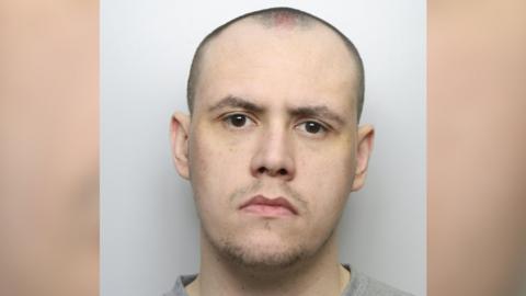 A police custody picture of Adam Martin. He has a shaved head and stubble and is wearing a grey jumper