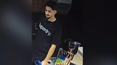A low quality image of a man wearing a black t-shirt with the words "Superdry" across his chest in white or silver lettering. He has black hair, and a beard across his upper lip and chin. He appears to be carrying some form of bag.