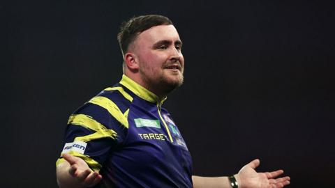 Luke Littler, in a purple and yellow top, reacts with his arms during his match against Rob Cross