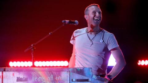 Coldplay lead singer Chris Martin on stage. He is wearing an off-white t-shirt and sitting behind a piano and microphone. A strip of red lights are visible behind him. 