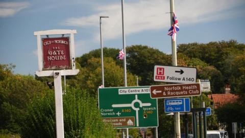A myriad of road signs and a pub sign approaching a roundabout near Norwich with three lamp posts each bearing a Union flag. Trees are seen in the distance.