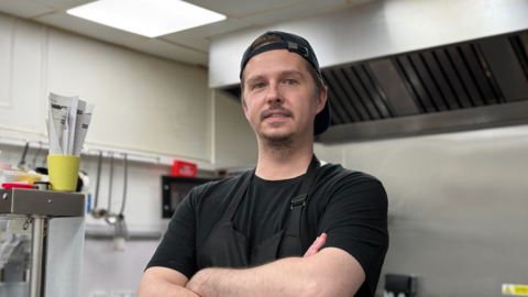 James Usher standing in the kitchen of The SeaGlass Cafe. He is wearing a black t-shirt and apron and a backwards baseball cap. There are ladles and an extractor fan behind him.