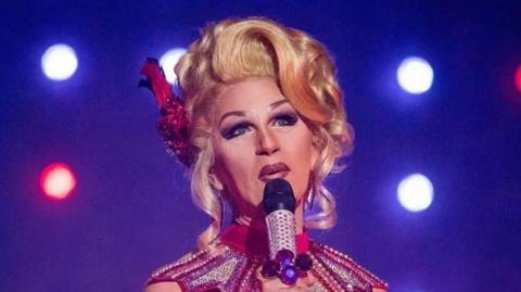 Head and shoulders shot of Zoe in full drag in a blonde wig, hair piled high and holding a microphone against a blue background with show spotlights