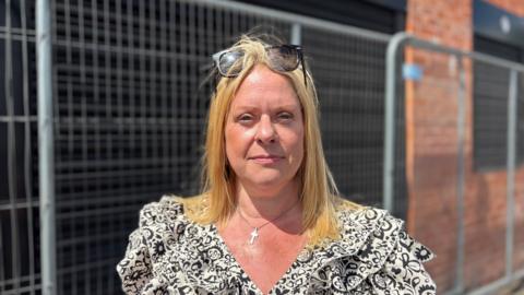 A woman with blonde straight hair and sunglasses on her head wearing a black and white summer dress stares at the camera not smiling in front of some metal railings