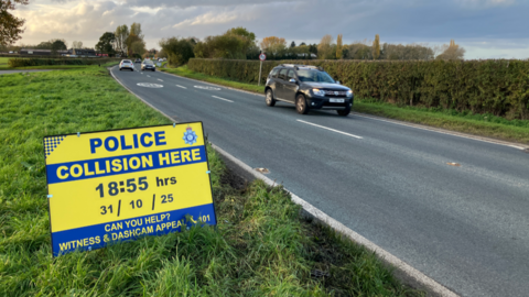 A view of a rural A-road. A sign in the foreground says "Police - collision here - 18:55 hours, 31/10/25.  Can you help? Witness & Dashcam appeal".  There are tyre marks in the grass verge.  The road behind has a sweeping bend. Several cars are passing.