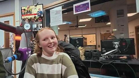 A head and shoulders shot of a smiling girl in a green and white striped jumper sitting in a radio studio, speaking into a purple Radio Kent microphone.