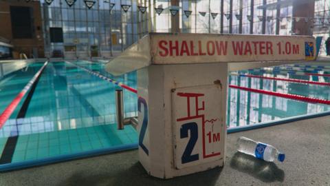 A indoor swimming pool with large glass windows behind it and a sign in the foreground which reads shallow water