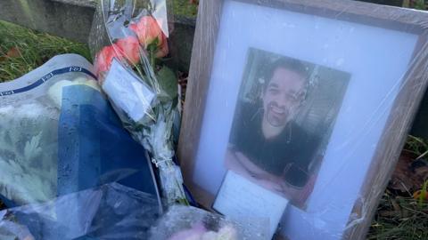 A framed image of Phil Tetley who was killed in a crash surrounded by bouquets of flowers next to a wooden fence
