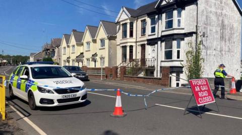 A police car is seen along a road, with police tape and a red "Road Closed" sign
