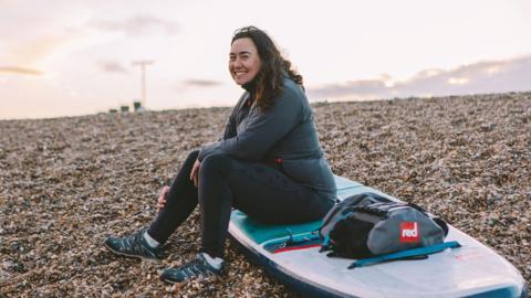 A woman with dark brown curly hair is sitting on a paddle board in sportswear. It is sunset and she is smiling as the paddleboard rests out of water on a stony beach.