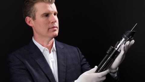 A man in a suit and wearing white gloves is carefully holding and admiring a lightsaber prop against a black background