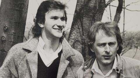 A black and white photo shows a taller man with dark shoulder length hair who is smiling and wearing a shirt, jumper and coat and a man with hair which is shoulder length at the back who is wearing a shirt and jacket. They are standing in front of two tree trunks.