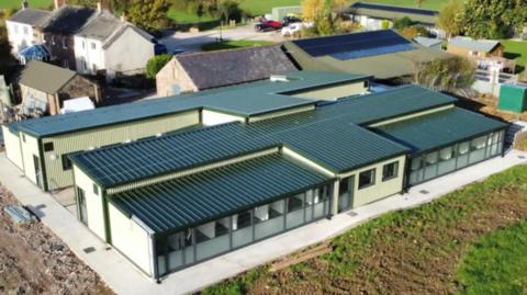 An aerial view of the new kennels - a large, white one-storey building with a flat blue ceiling. It is surrounded by several other buildings and green fields.