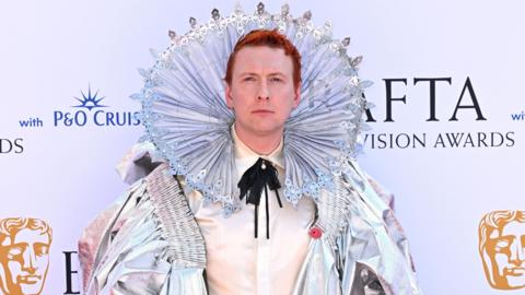 A man with a shiny outfit on looking into the camera and smiling. He has red hair and he has a metallic ring around his face/ head. 
