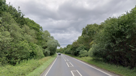 A general view of the A43 in Northamptonshire. Two cars can be seen in the distance. The road is lined with trees and bushes.