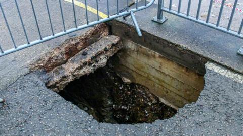 A large sinkhole in the road of a seafront promenade. It reveals a void underneath the surface. It is fenced off with silver-coloured fences. 