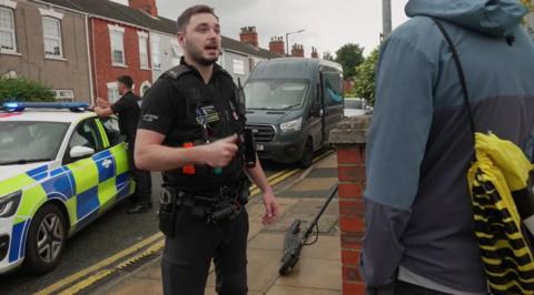 A police officer talking to young person in the street, with an e-scooter propped up against a wall. There is a police car and a second officer in the background.