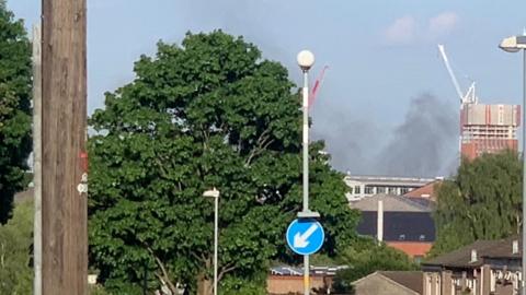 A thick plume of smoke in the sky above buildings and trees.