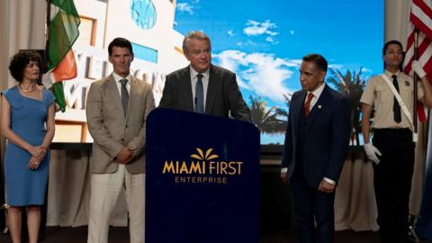 A formal press or announcement event taking place indoors, staged with a podium and a branded backdrop.