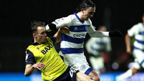 An Oxford United player in yellow tussles for the ball with a QPR player in blue and white hoops