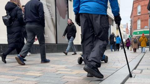 People walking on a busy high street, wearing coats an hats on a clear day in Douglas city centre.