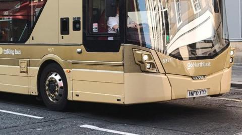The front of a gold Go North East bus on the road. The driver is wearing a white shirt but their face is obscured by the vehicle's window frame.