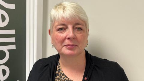 A woman with short hair looks at the camera. She is wearing a black cardigan and a shirt with leopard print.