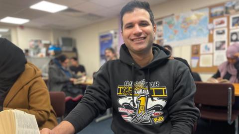 A smiling man in a black hoodie sits on an office chair with a map of the World behind him.