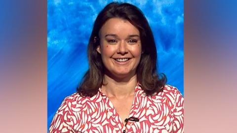 Gillian Smart has shoulder length dark hair, wears a patterned shirt and jeans