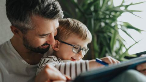 A young boy sitting on a man's knee. They are sitting in a home and drawing on a tablet. They are both smiling.