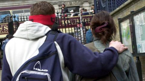 A male and female teenager stand with their backs to the camera. The male is to the left of the female with his arm across her back holding her shoulder. The woman is wearing a denim jacket while the man is wearing an oversize navy/white sports hoodie. 