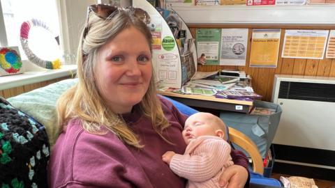 A smiling woman with blonde hair is holding her sleeping baby in her arms while sitting on a sofa. She is in a community building, with lots of leaflets put up on the wall behind her.