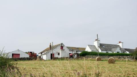 A view of a farm from a distance with white buildings, hay bales in a field and grey skies above