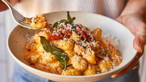 A person holds a white bowl in one hand and a fork in the other. In the bowl is golden gnocchi topped with parmesan cheese and green leaves