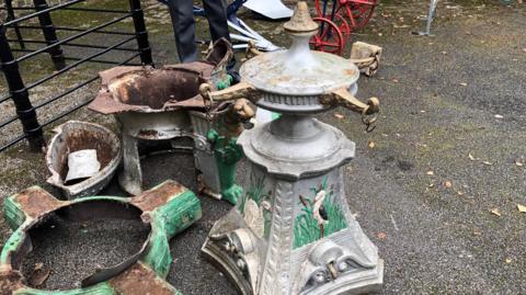 A heavy cast iron plinth painted silver with depictions of a swan on one side and a black and white water bird on the other. Both have reeds and bull rushes in the background, the object stands about 1m (3ft) high and has four gold arms, about 15cm (6 inches) in length coming off a circular shaped piece. Rings are attached to each arm, which would have originally had a chain on attached to a cup to collect drinking water. The large base, which is damaged, rusted and painted green, is next to it.