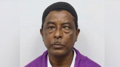 Headshot of a man looking straight ahead with a small black cap on his head, wearing a magenta shirt