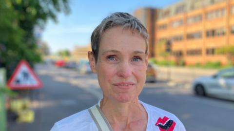 Katie, a woman with short golden hair. She is wearing a white T-shirt and smiling for the camera.