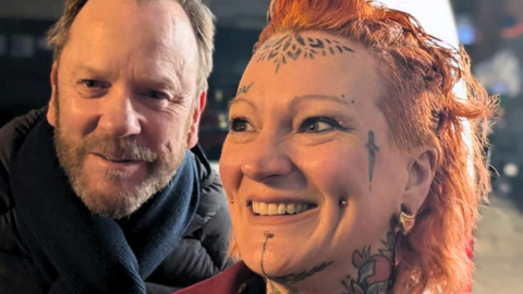 A tattooed woman with orange hair smiles next to Kiefer Sutherland, a bearded man in a black jacket.