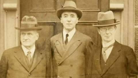 A black and white archive photo of Joseph Fisher, Richard Feetham and Eoin MacNeill during a meeting of the Boundary Commission in Armagh in 1924.  They are all wearing suits. ties and hats and are standing in front of a door.