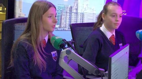 Two girls in school uniforms are in a radio studio in front of microphones.