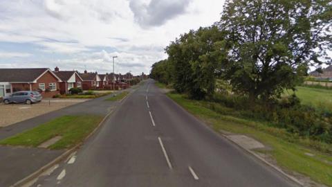 A road with residential houses on the left and trees on the right. The sky is overcast.