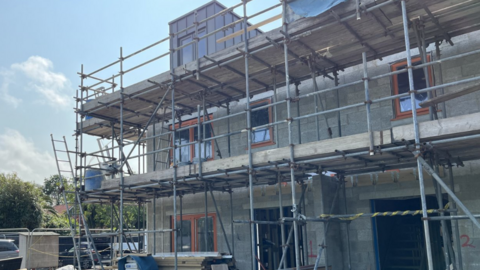The image shows a construction site where a building is in the process of being constructed. Scaffolding surrounds the partially built structure, and several windows have already been installed. Various construction materials—such as wooden planks, metal rods, and machinery—are scattered around the site. 