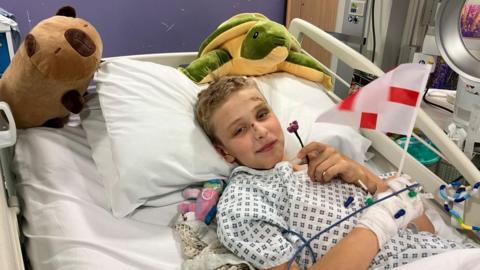 Martin smiles at the camera as he lies in his hospital bed. Wearing a hospital gown he holds a small England flag in his heavily bandaged right had and a crocheted rose in his left hand, which was given to him by the players. There are two cuddly toys resting on either side of his pillows, a green turtle and a brown dog