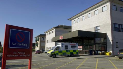 An ambulance is parked outside the entrance of a hospital building. A red no parking sign is on display, it says no parking, helicopter landing pad. The hospital building is grey. 