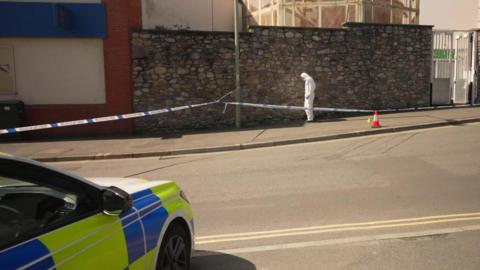 A police forensics officer inspects the scene of a double stabbing in Newton Abbot. The officer is stood within a taped off area on a street and is wearing white overalls. A police car is in the foreground.