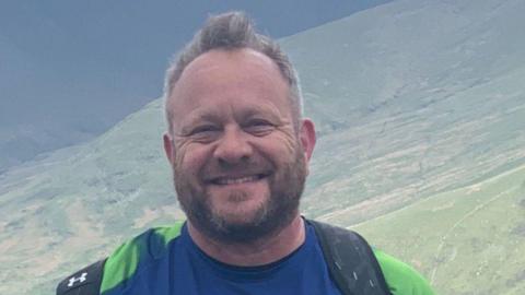 A man smiles into the camera, with green hills in the background