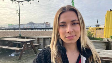 A 25-year-old woman with long blonde hair. She is looking at the camera and wearing a black fleece. She is stood on a decked area with wooden picnic tables around her. The decking is in front of Brighton Palace Pier, a white pier, which can be seen in the left background of the image. The sky is cloudy