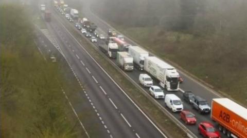 Queuing vehicles, including cars and lorries on a road in slightly foggy conditions.