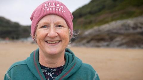 Carol Maddock stands on a beach smiling at the camera. She is wearing a green hoodie and a pink beanie hat that says "ocean therapy" 