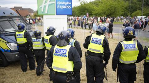 A picture from behind a line of riot police officers being confronted by an angry crowd outside a Holiday Inn Express in Rotherham